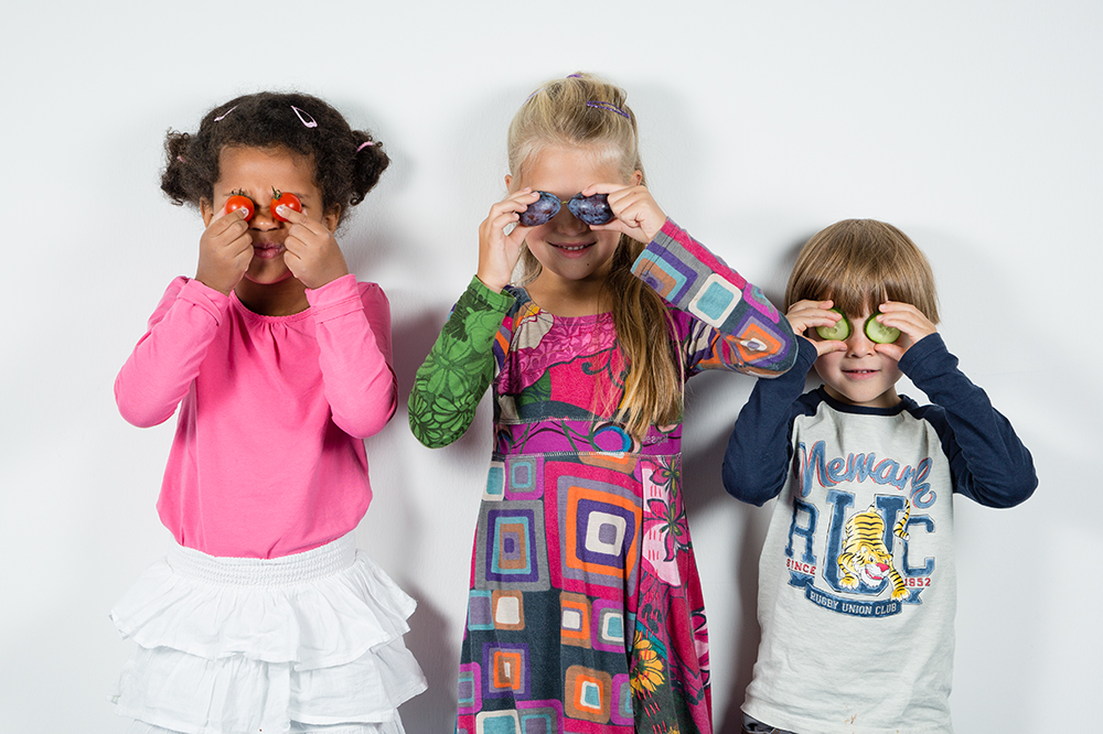 Three children holding vegetables and fruits in front of their eyes.
